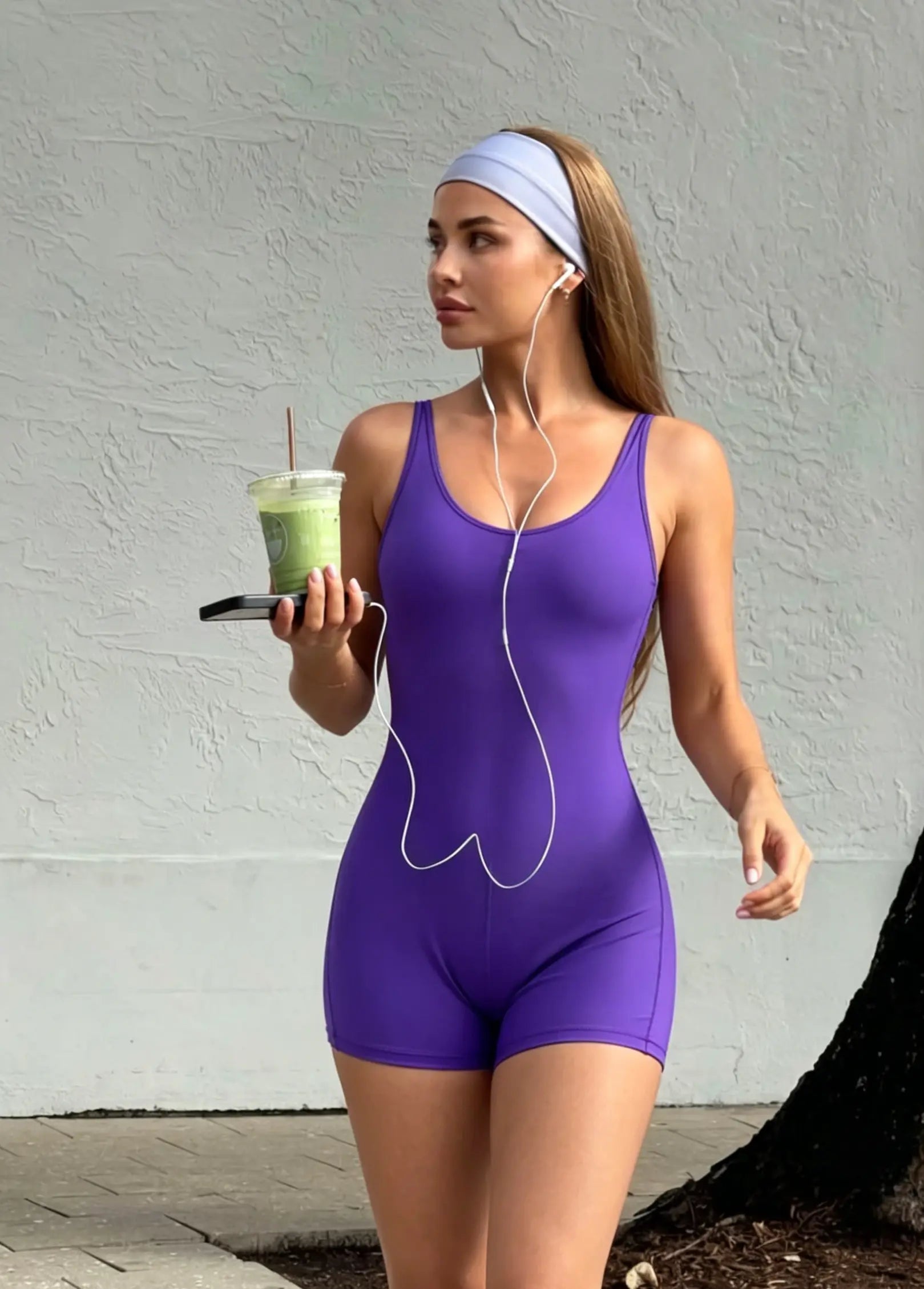 Woman in a purple bodysuit holding a green smoothie against a gray wall.