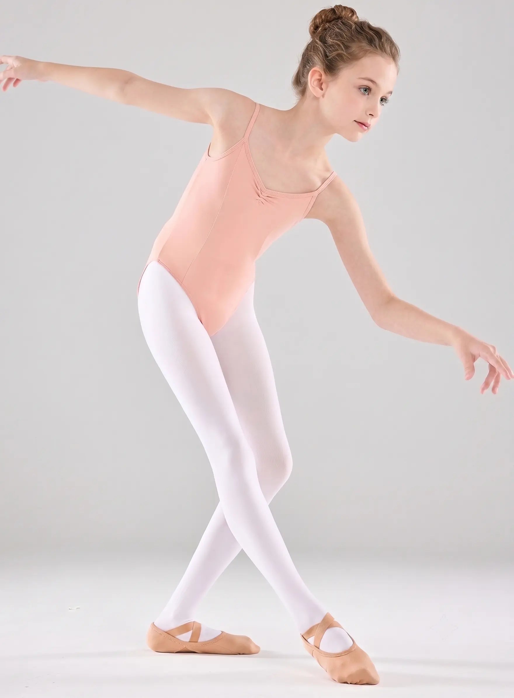 A young girl dancer in a ballet pose, wearing a light peach pink leotard, white tights, and tan pointe shoes against a light gray studio background.