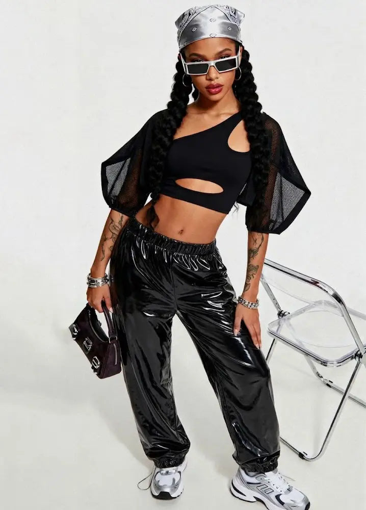 Person wearing a black outfit with sheer sleeves and shiny pants, holding a black handbag on a white background.